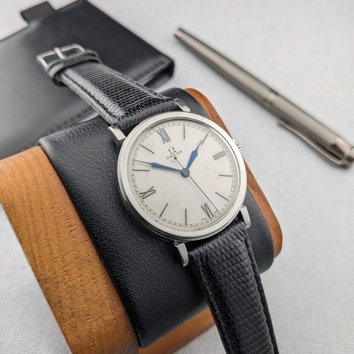 Omega watch resting on a black-and-wood cushion, with a black wallet and silver pen arranged behind it.
