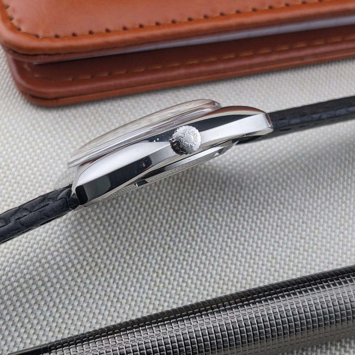 Side profile of the Longines Ultrachron reference 8429 showing its ridged crown, curved case edge, crystal, black strap and silver pen.