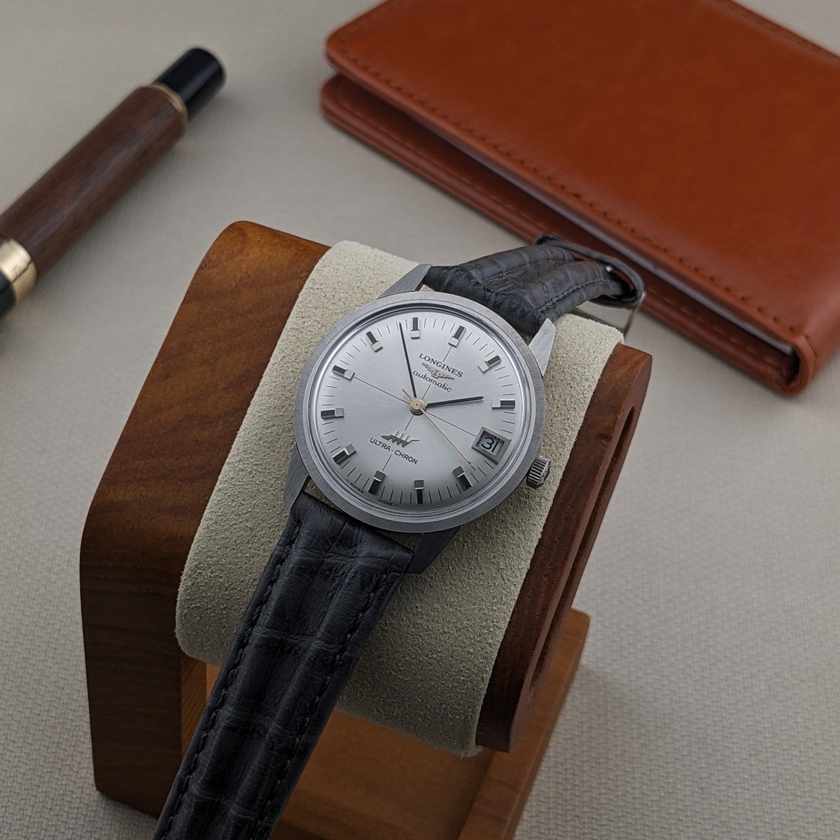 Longines Ultra-Chron 7827 centered on a wood-and-fabric watch cushion, viewed from above with the date window and black strap visible.