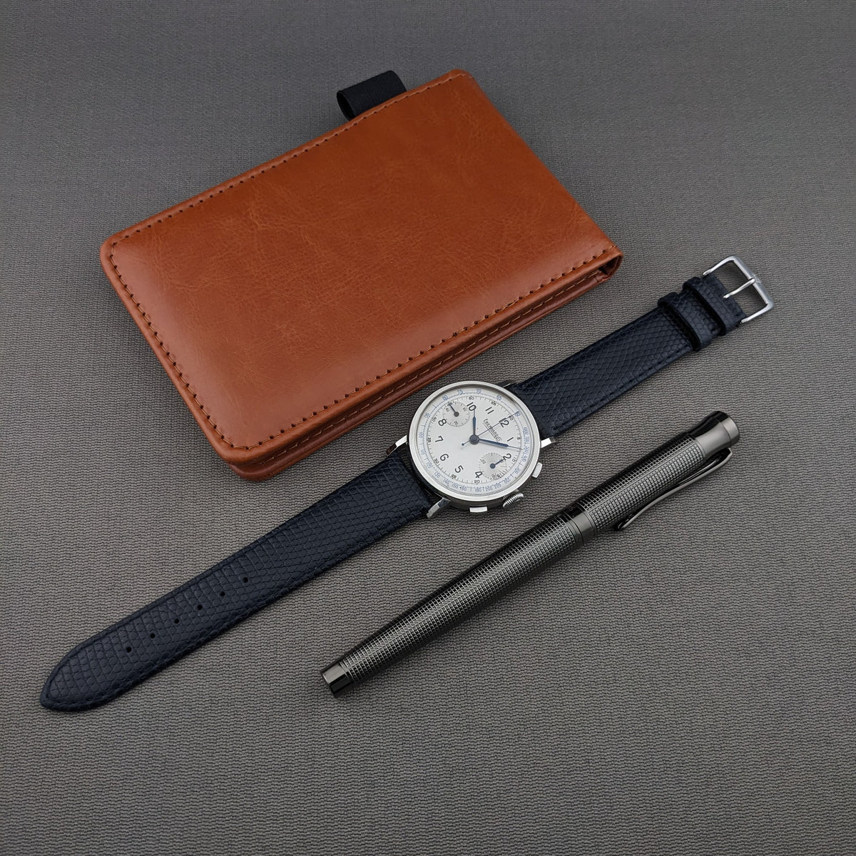 Eberhard & Co.. Pre-Extra Fort with blue strap is arranged beside a brown wallet and textured metal pen on gray fabric.