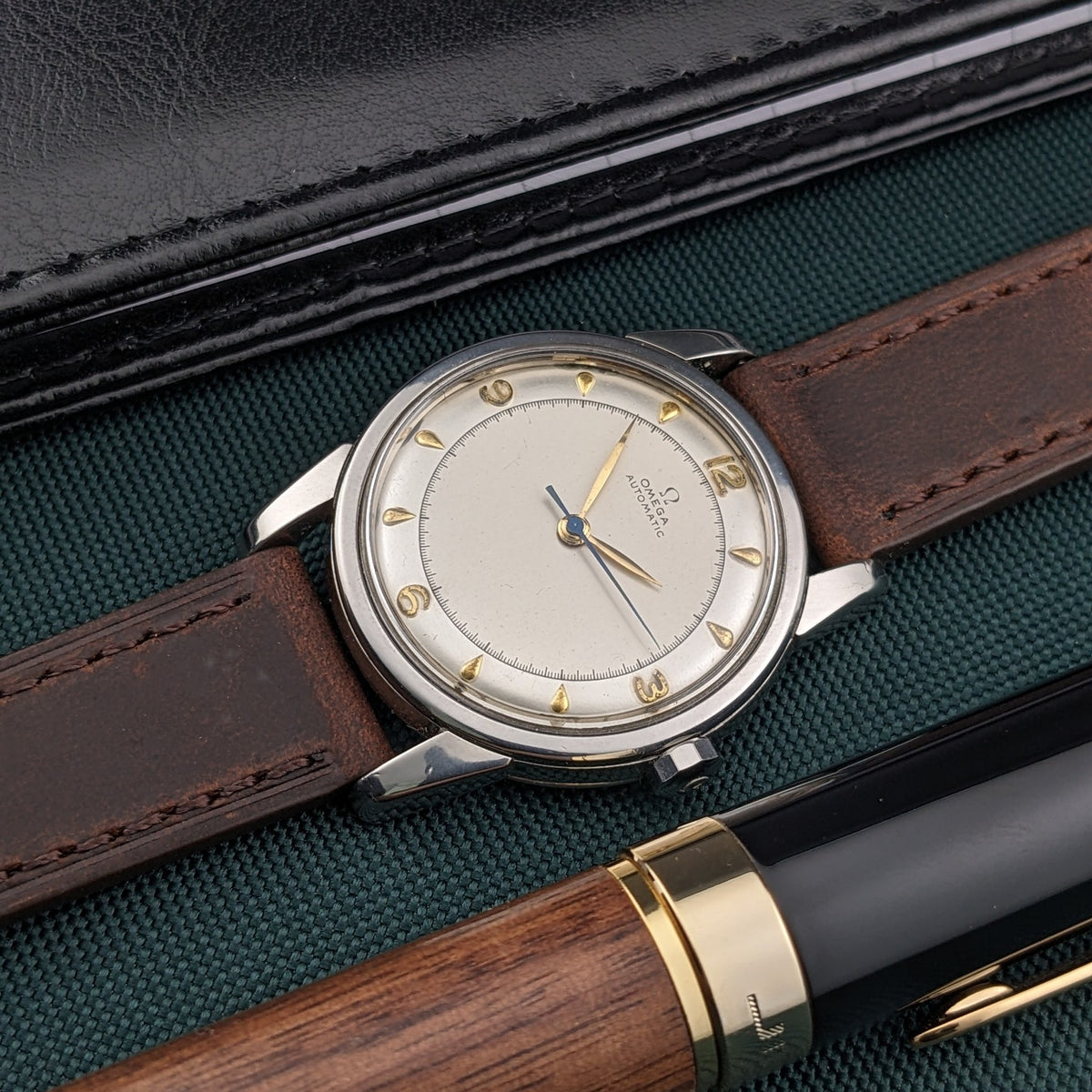 Omega Automatic Bumper reference 2767-4 SC rests diagonally on green fabric beside a wood-and-black pen and a black leather case.