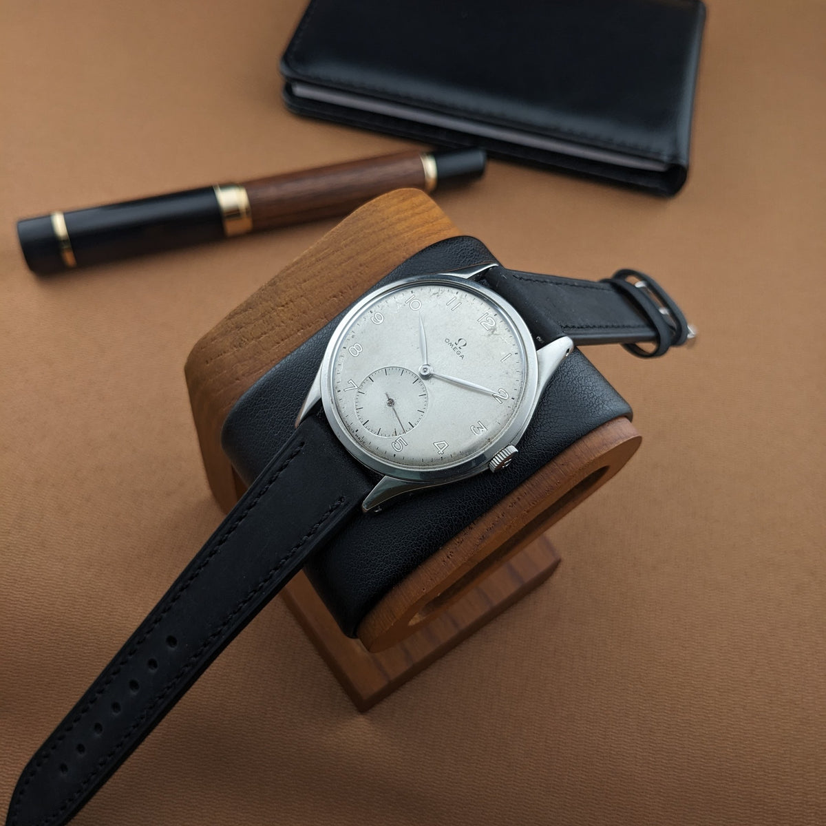 Omega Jumbo reference 2505-5 resting on a wooden watch stand with its black strap extended, alongside a pen and black wallet.