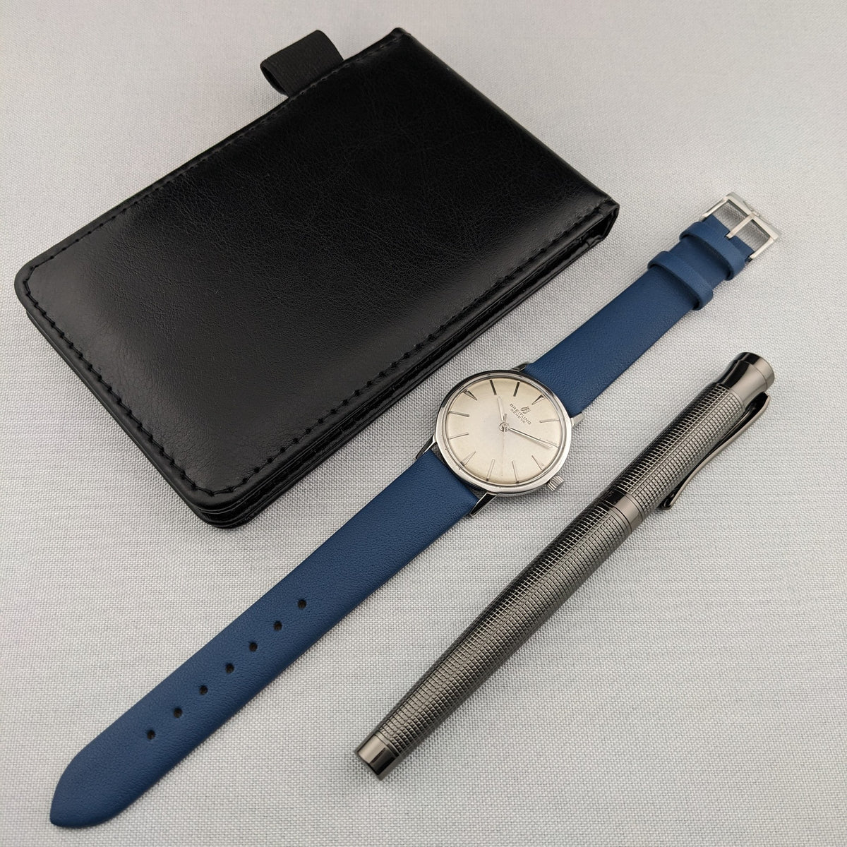 Breitling Automatic watch with blue strap shown beside a black wallet-style case and a silver textured pen.