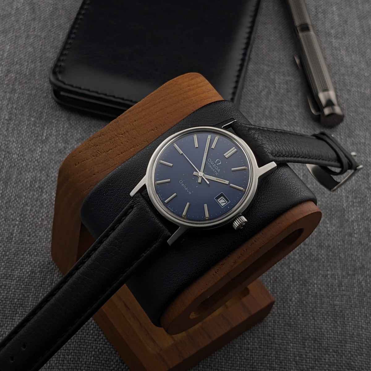 Omega Geneve Automatic reference 166.0163 arranged on a wooden watch stand, with blue dial, black strap and surrounding desk accessories.