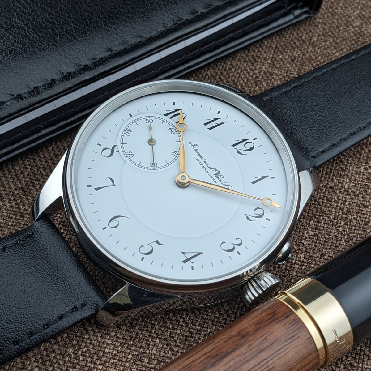 IWC Schaffhausen डायल का कोणीय फोटो, काले चमड़े के केस और लकड़ी के लेखन उपकरण के साथ, सुनहरे हाथ और सेकंड्स सबडायल दिखाता हुआ।