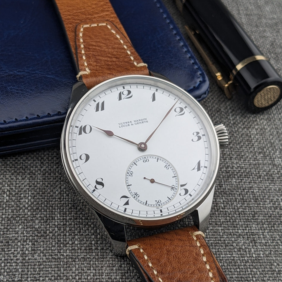 Ulysse Nardin Locle & Genève घड़ी नीले चमड़े के केस और काले लेखन उपकरणों के साथ प्रदर्शित, डायल ऊपर की ओर।