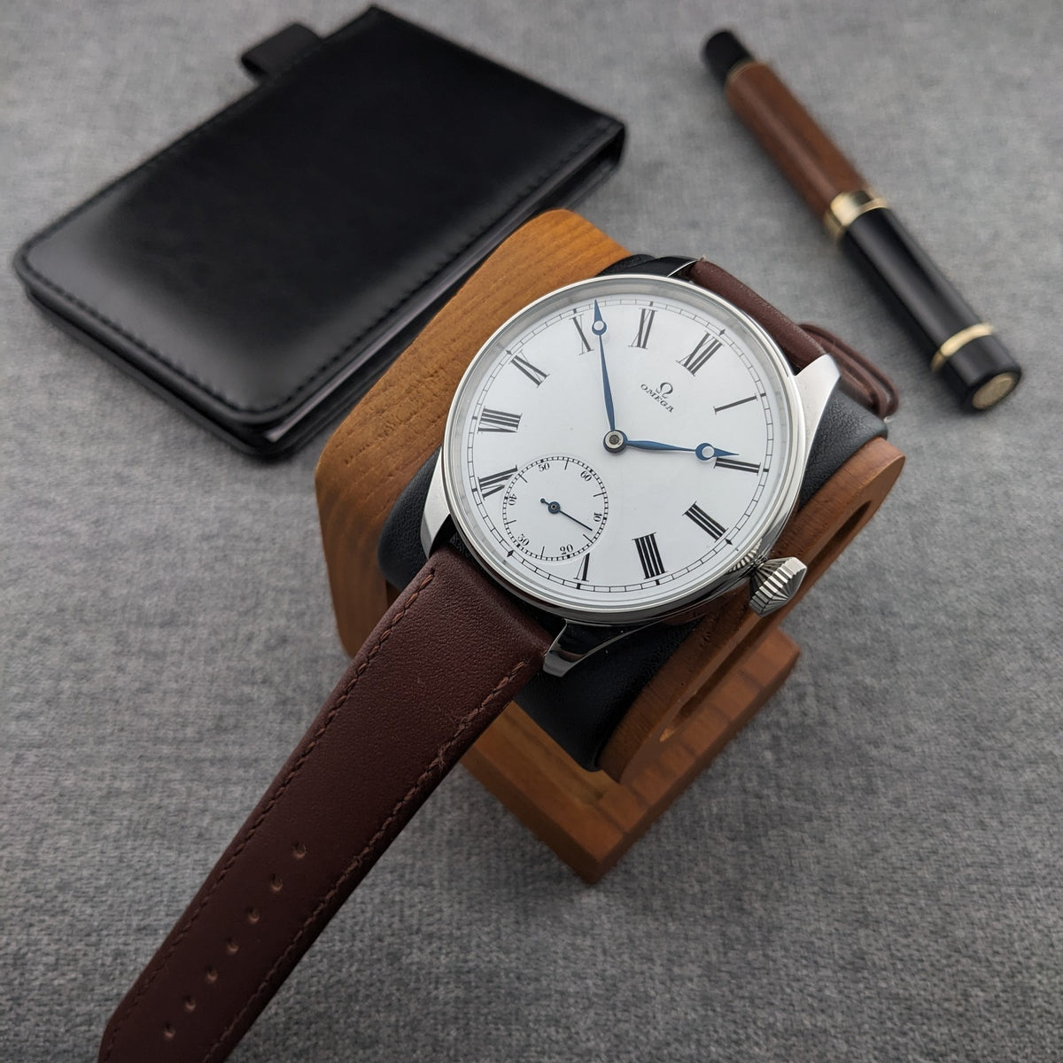 Orologio Omega su supporto in legno con cinturino marrone esteso, accanto a una custodia nera e una penna in legno e nero.