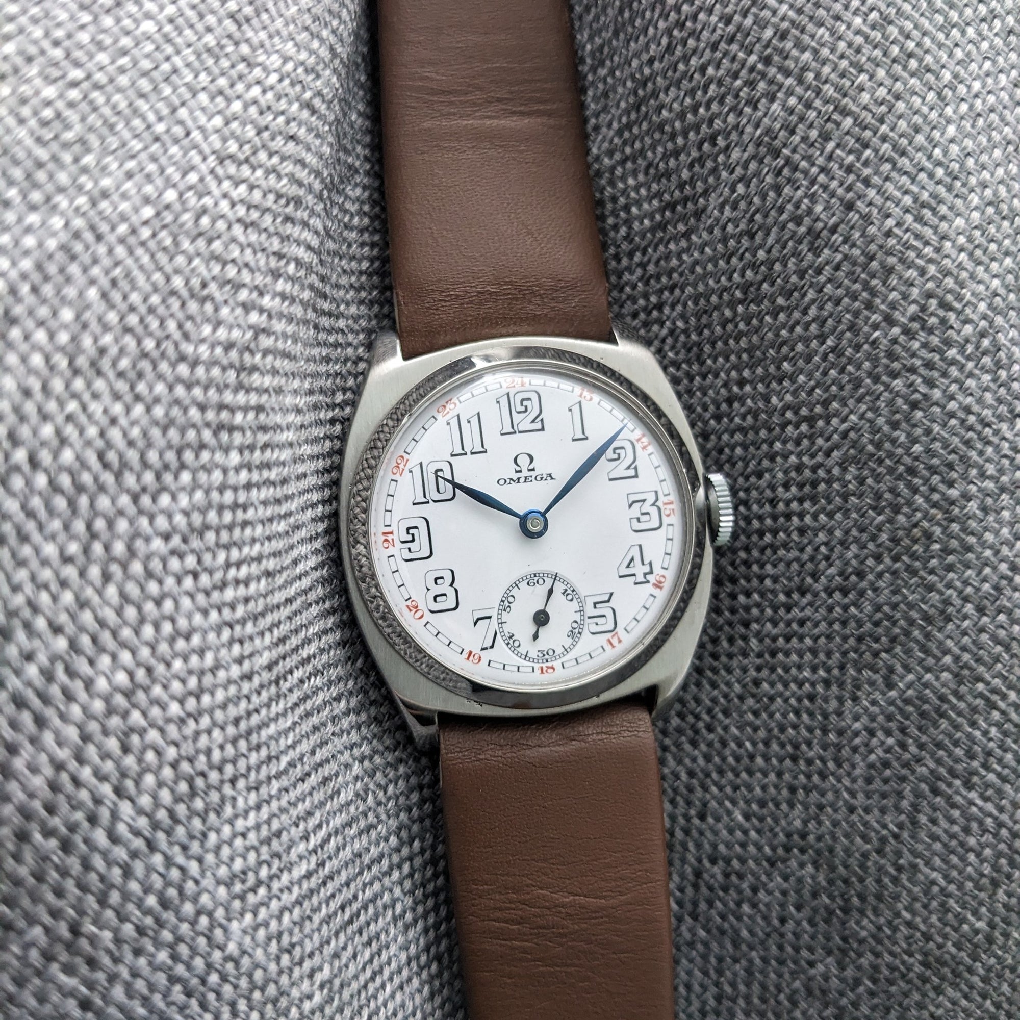 Omega watch presented vertically on gray fabric, showing its white dial, blue hands, Arabic numerals, crown, and brown leather strap.