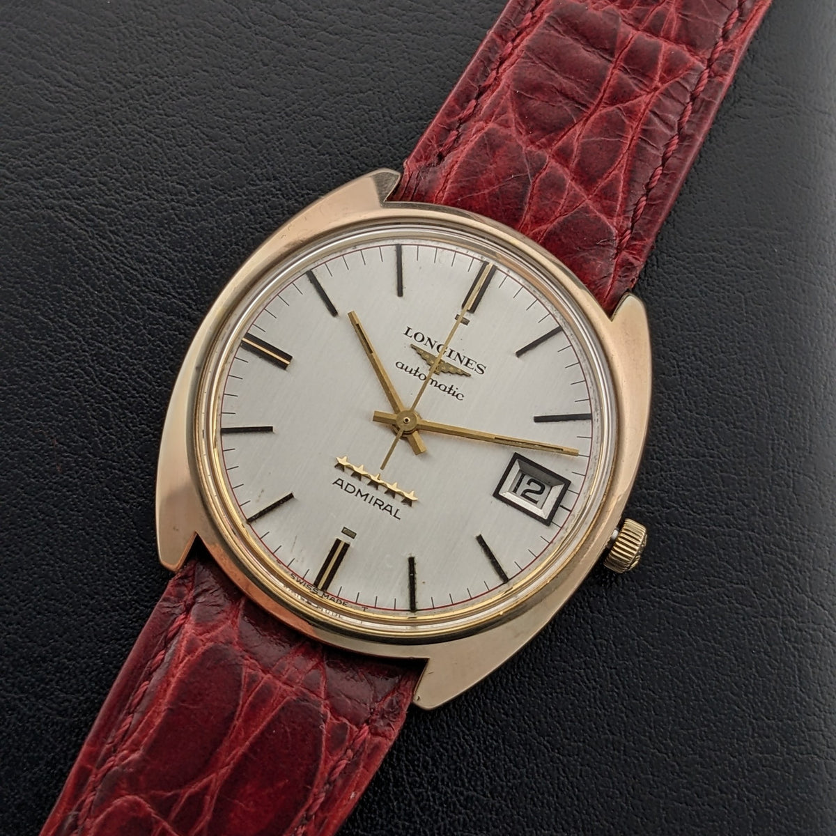 Longines Admiral 8343-1 vor schwarzem Hintergrund, hebt silbernes Zifferblatt, goldfarbenes Gehäuse, Datumsfenster und burgunderrotes Armband hervor.