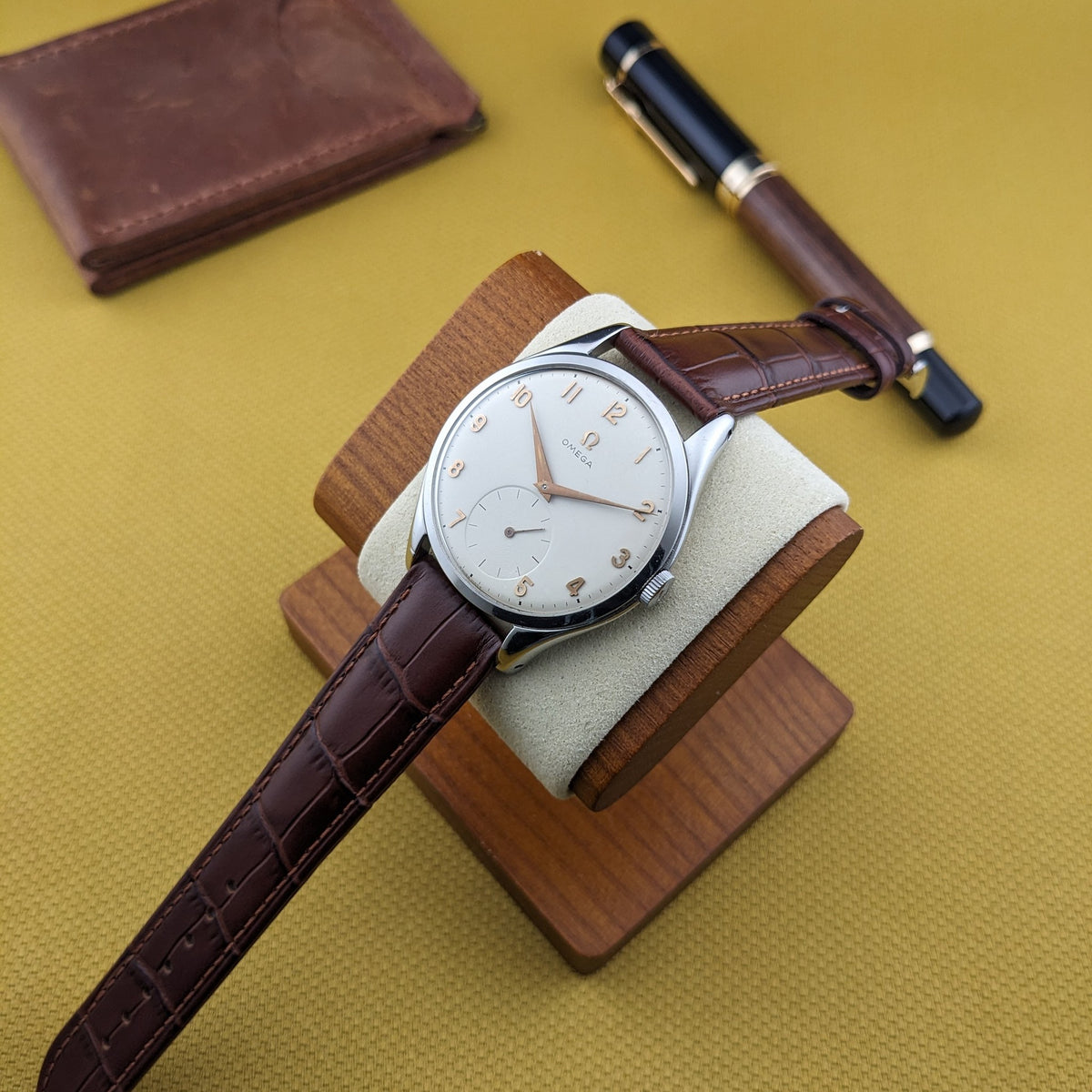 Omega Jumbo Referenz 2505-10 auf einem Holz-Uhrenständer, mit braunem Portemonnaie und Stift im Hintergrund arrangiert.