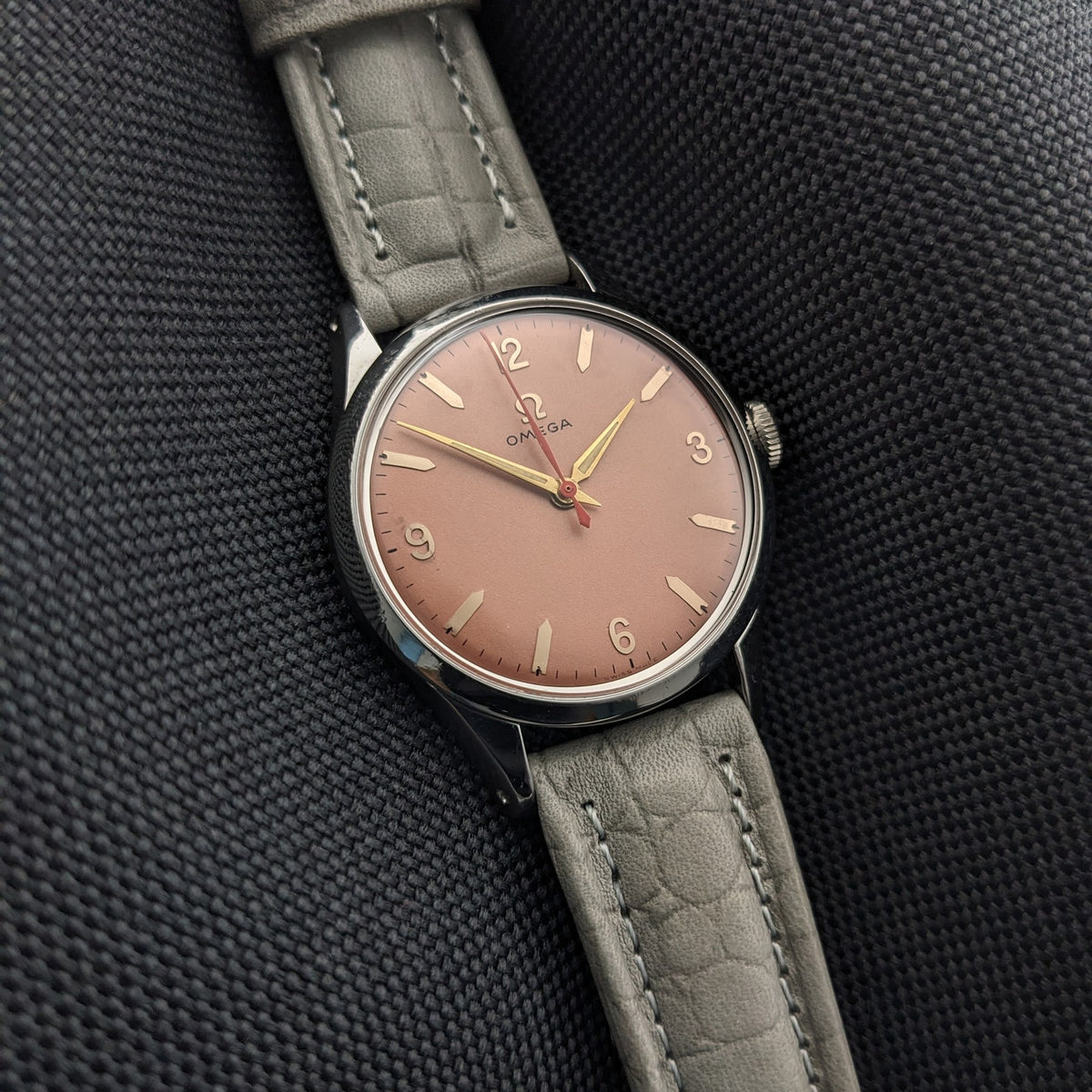 Omega Classic Referenz 2760-6 SC aus einem Winkel auf schwarzem Stoff, hebt das rosafarbene Zifferblatt, poliertes Gehäuse, Krone und graues Armband hervor.