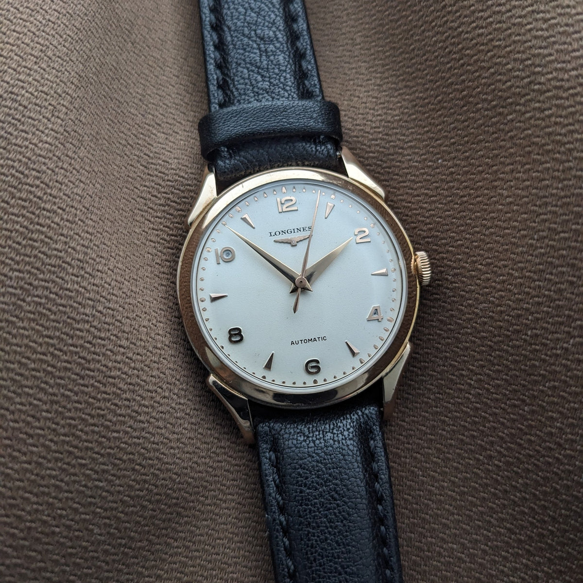 Frontansicht der Longines Automatik Referenz 6058 auf braunem Stoff, zeigt ihr helles Zifferblatt, Markierungen, Zeiger und schwarzes Lederarmband.