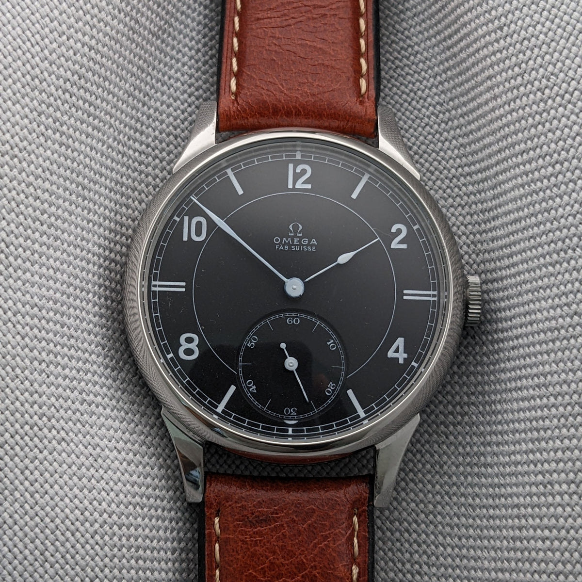 Nahaufnahme eines Omega-Zifferblatts mit arabischen Ziffern, zentralen Zeigern, Minutenanzeige und kleiner Sekunde auf grauem Stoff.