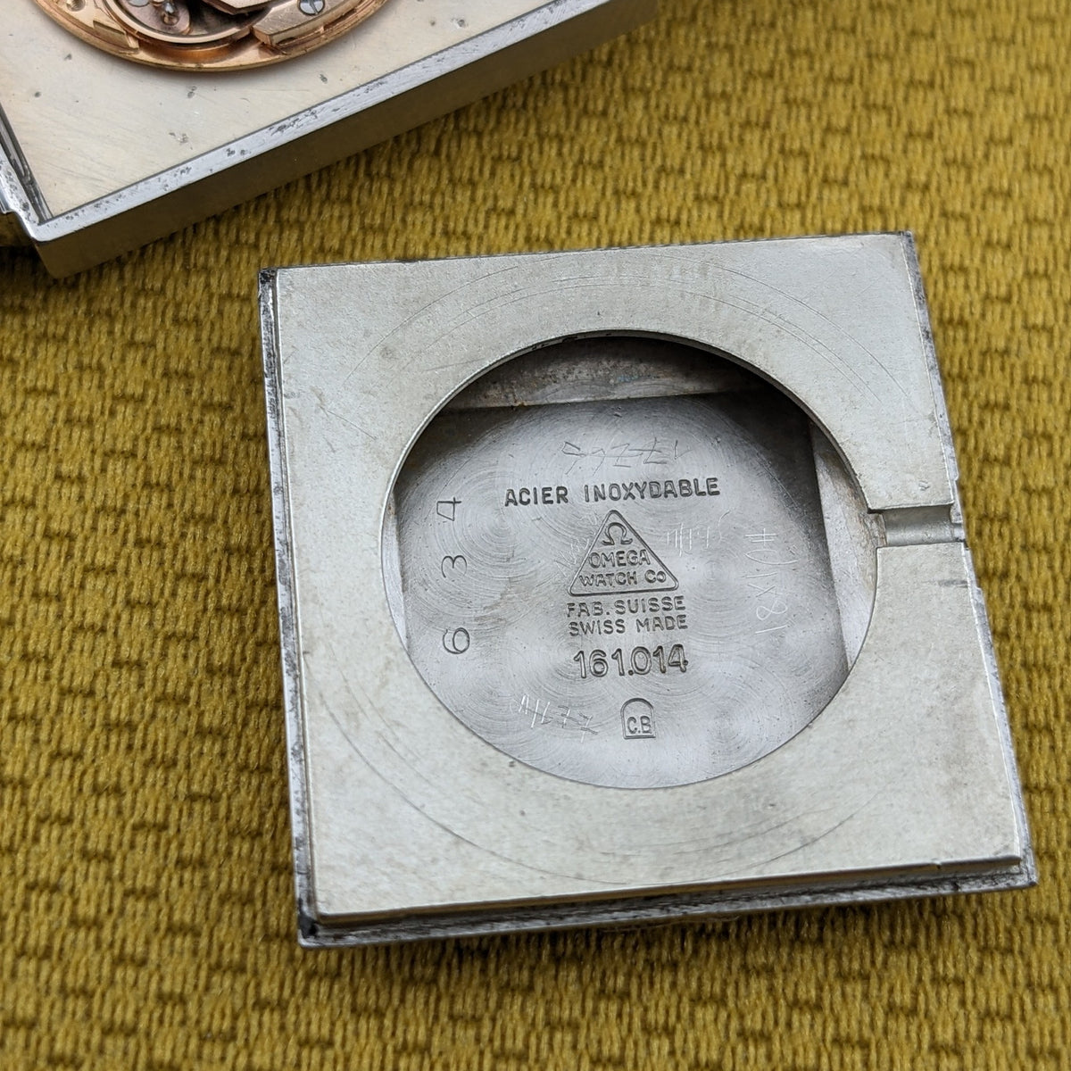 Omega De Ville Automatik 161.014 Gehäuseboden neben der Uhr, mit sichtbarem graviertem Text und kreisförmig vertieftem Zentrum auf senffarbenem Stoff.