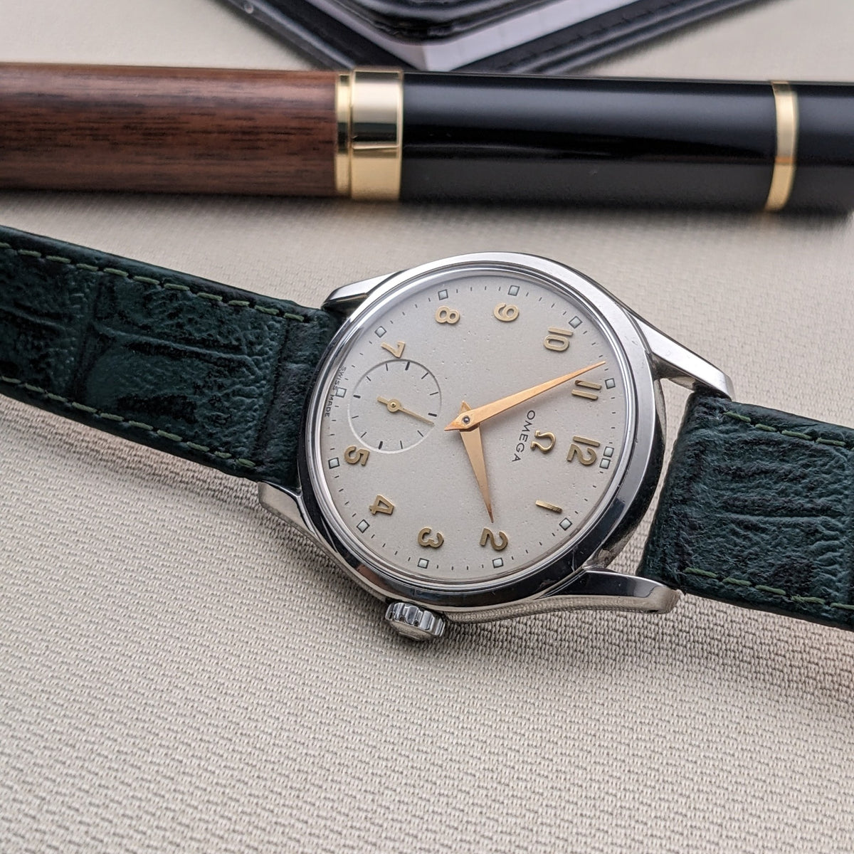 OMEGA Classic 2639-11 esposto orizzontalmente con quadrante argentato, lancette dorate, cinturino verde e penna in legno e nero sullo sfondo.