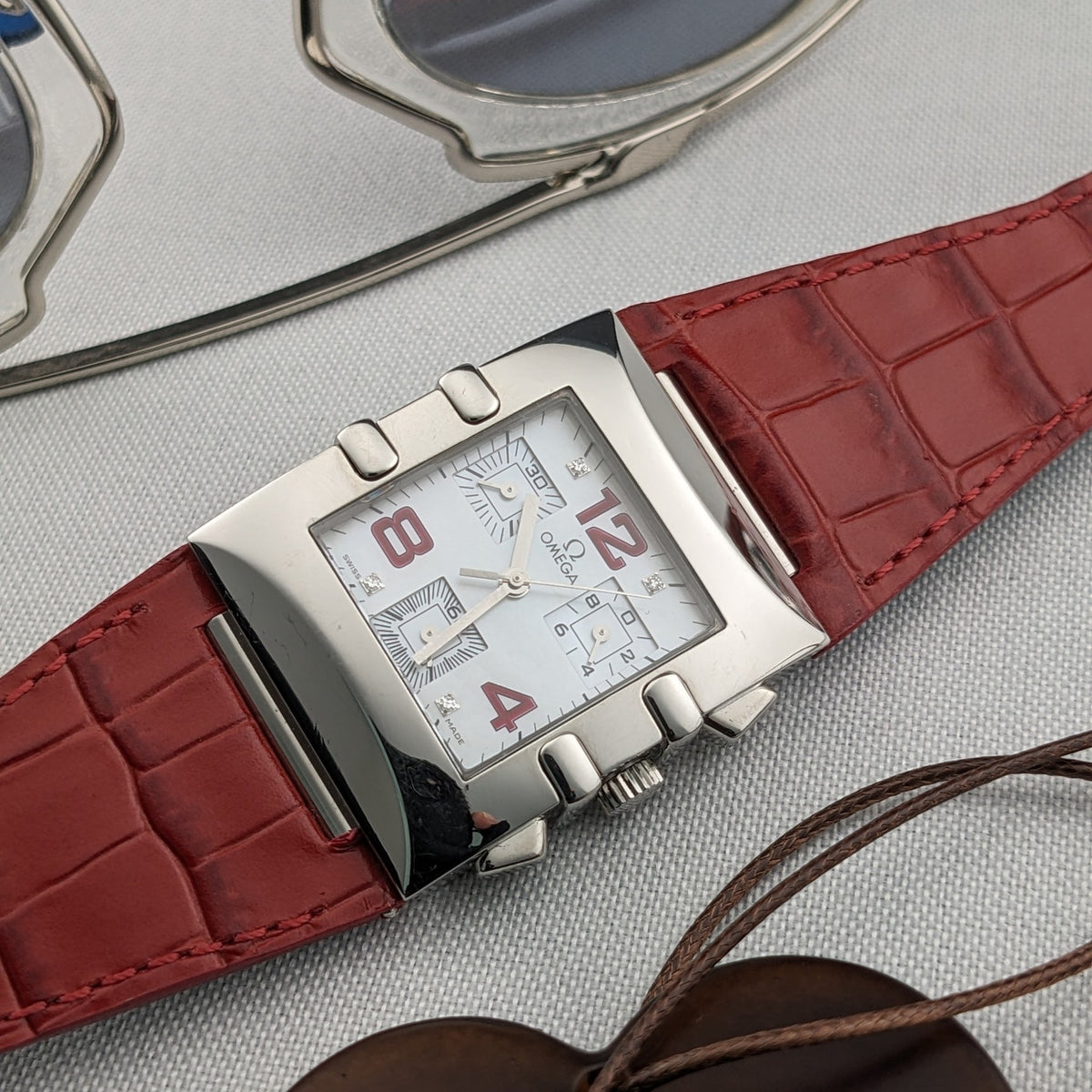 Omega Constellation Quadra 1841.73.31 at an angle with a white dial, red numerals, rectangular case, red strap, and pendant cord nearby.