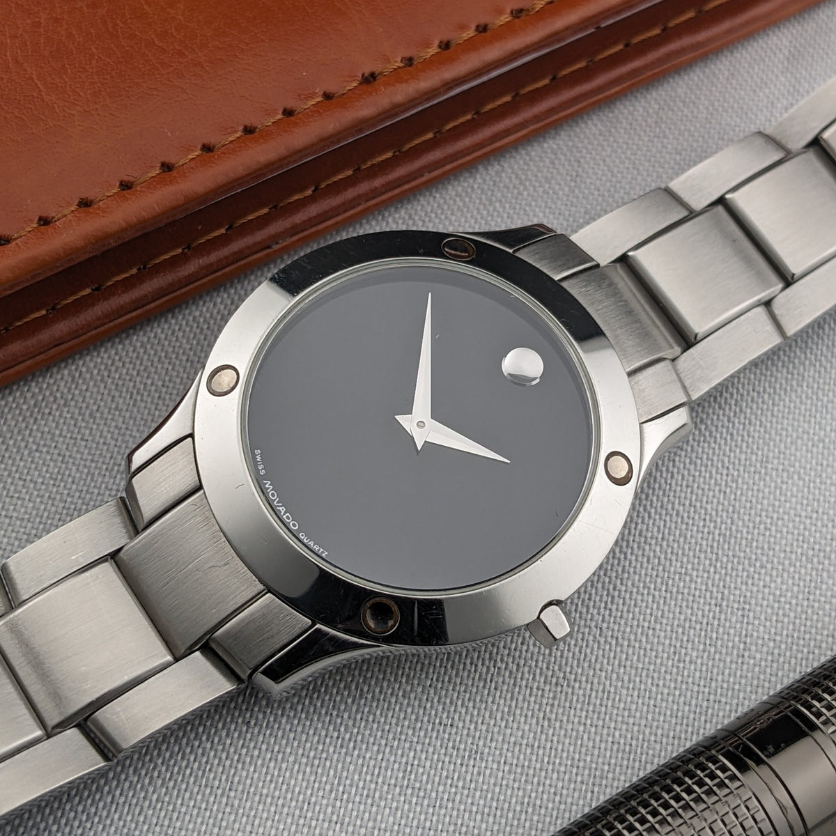 Vue rapprochée du cadran noir Movado avec aiguilles argentées, repère circulaire, lunette polie et maillons de bracelet adjacents.