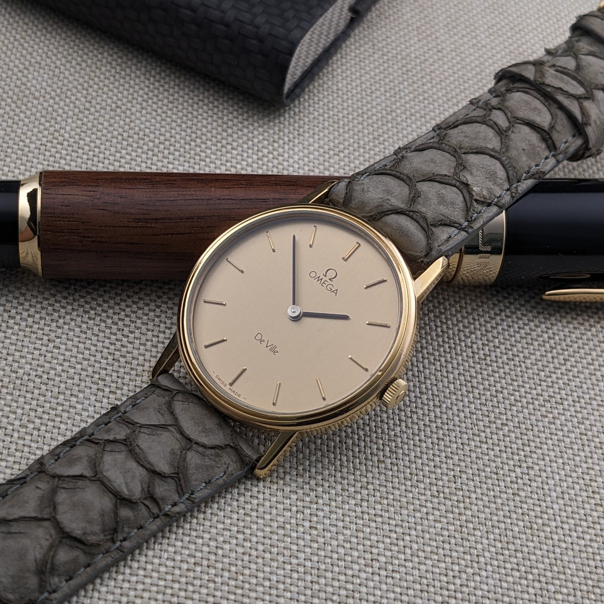 Omega De Ville reference 111 0156 resting diagonally on a gray strap, with a wood-and-black pen and dark case in the background.