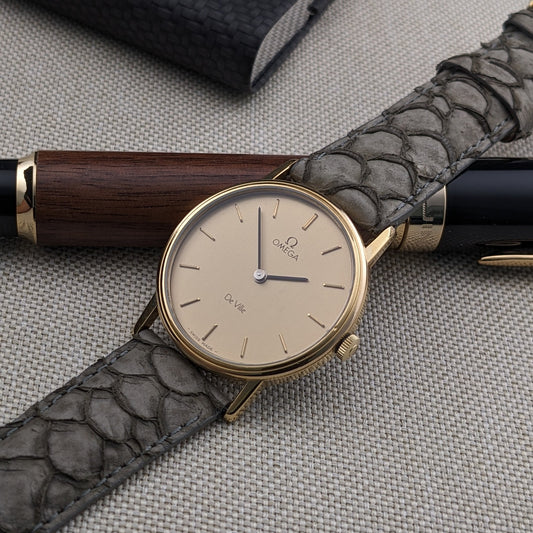 Omega De Ville reference 111 0156 resting diagonally on a gray strap, with a wood-and-black pen and dark case in the background.
