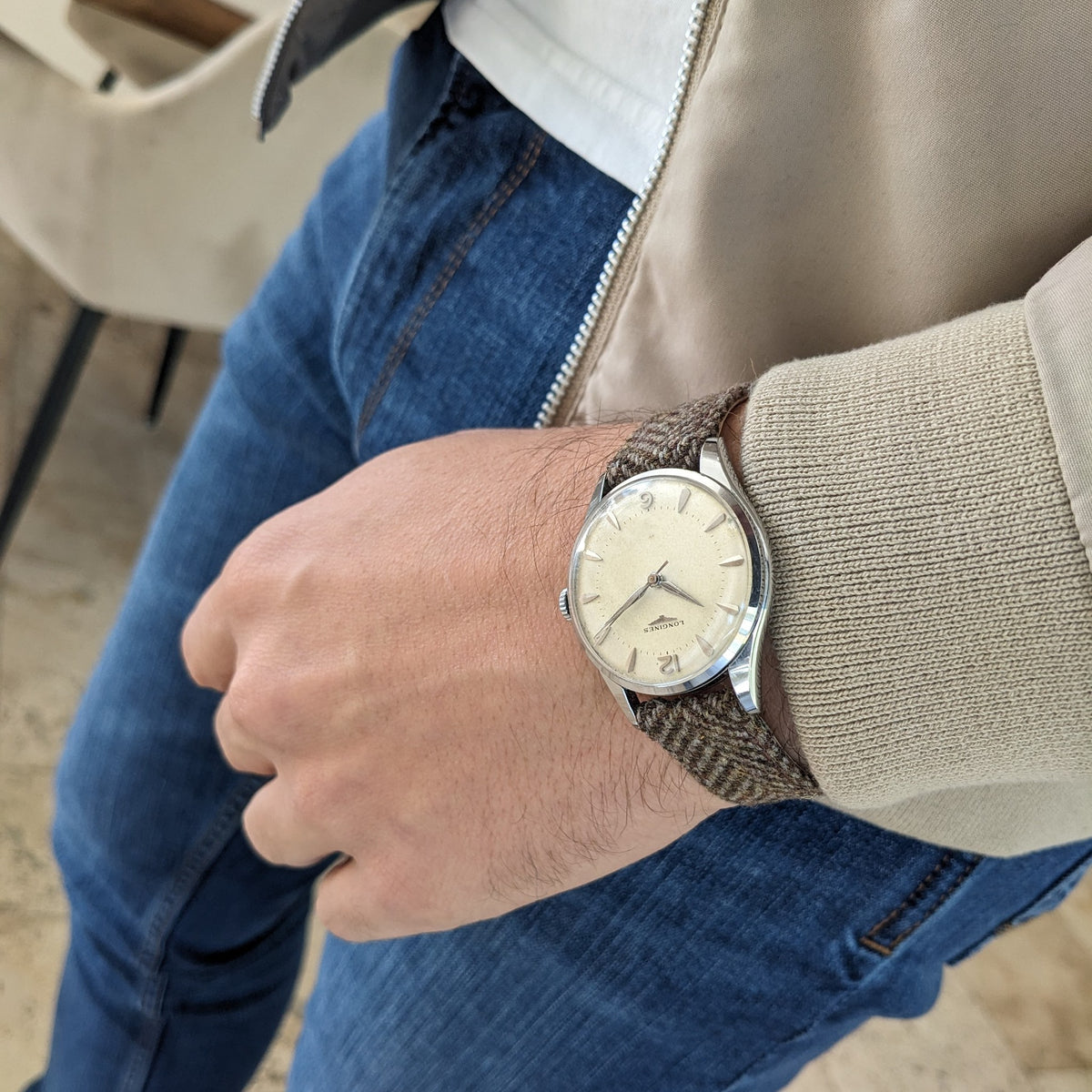 Longines Classic reference 8135-5 worn on a seated wrist, with its cream dial and brown woven strap visible over a beige jacket and blue jeans.