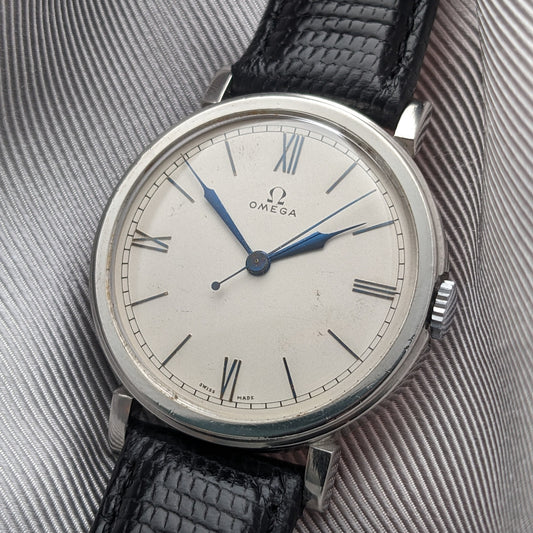 Close view of the Omega dial with Roman numerals, blue hands, minute track and black textured strap against gray fabric.