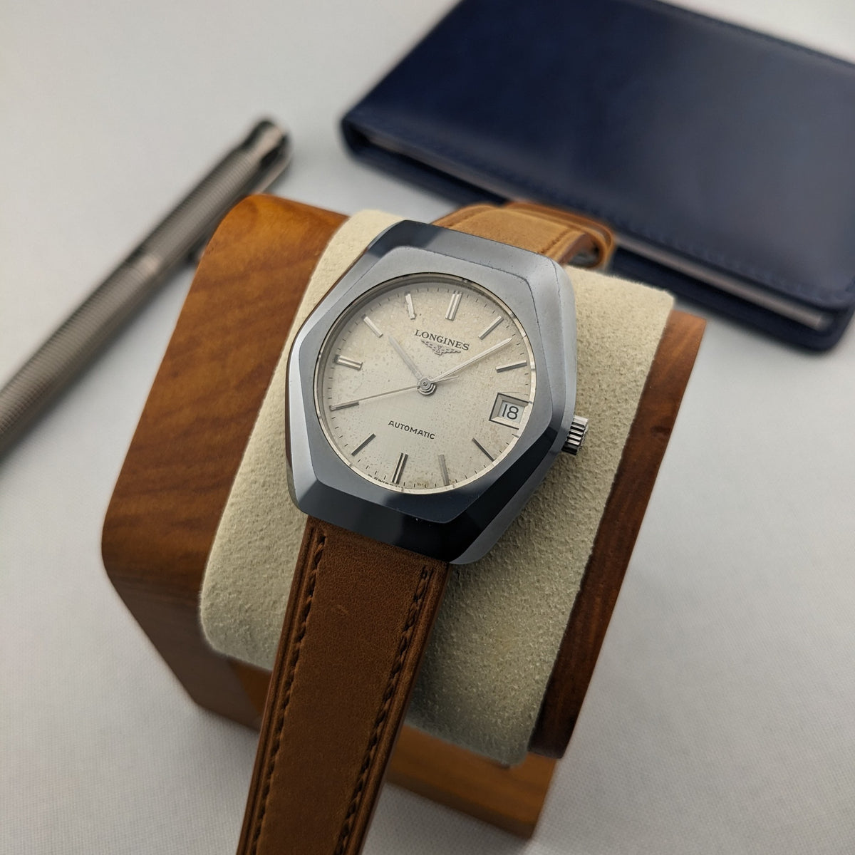 Angled front view of the LONGINES Automatic reference 3042-633 on a wooden stand, with cream dial, brown strap and blue case behind.