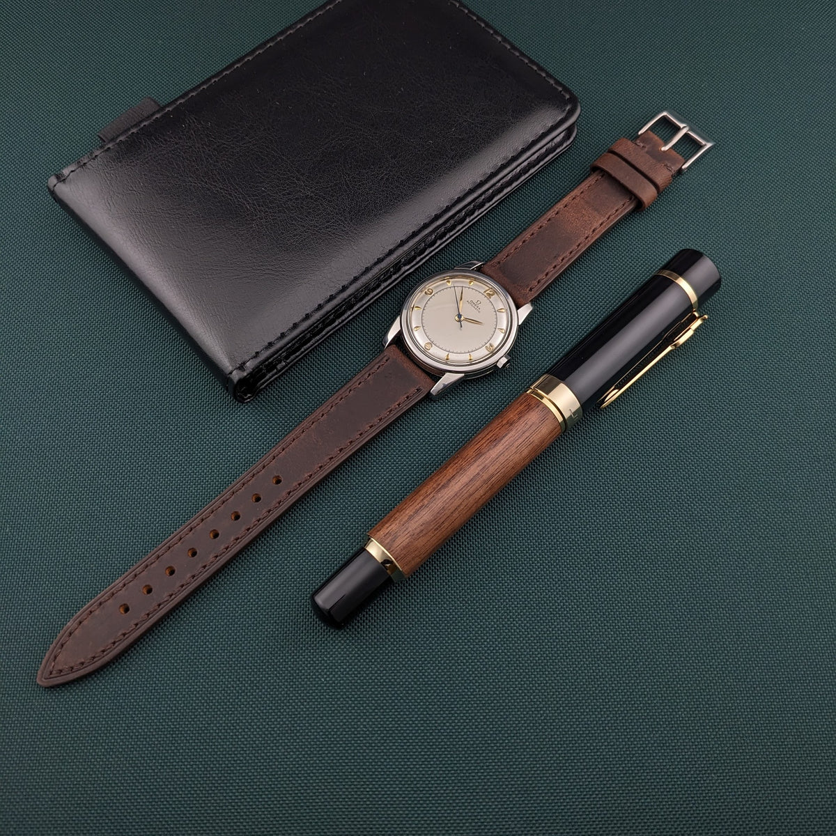 Omega Automatic Bumper reference 2767-4 SC, brown leather strap, black leather case and wood-and-black pen arranged on green fabric.