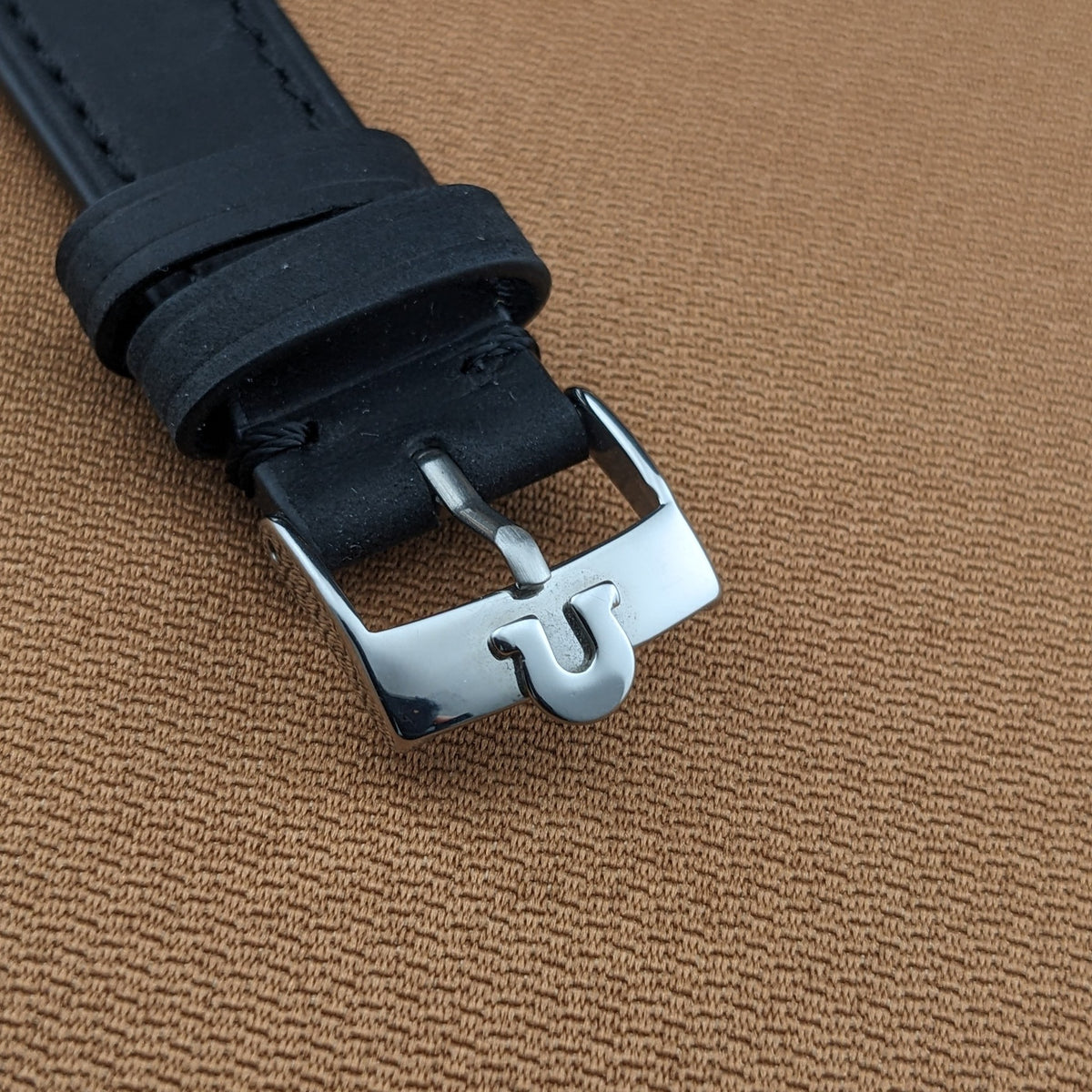 Close-up of the black leather strap and polished Omega buckle on the Jumbo reference 2505-5, set against brown fabric.