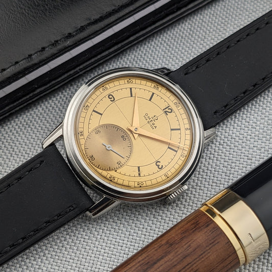 Reloj Omega Sector con esfera crema y correa negra junto a una pluma fuente de madera y negro sobre tela gris.