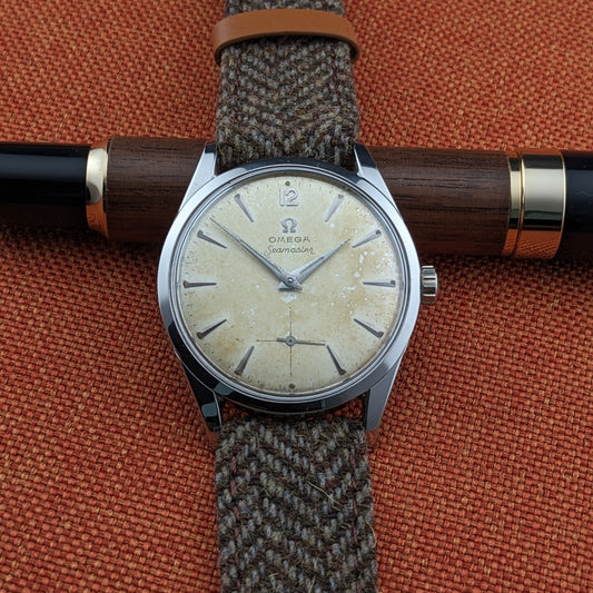 Omega Seamaster रेफरेंस 29374 लकड़ी और काले पेन के ऊपर रखा हुआ, क्रीम डायल, घंटे के मार्कर्स, हाथ, सेकंड्स सबडायल और बुना हुआ स्ट्रैप दिखा रहा है।