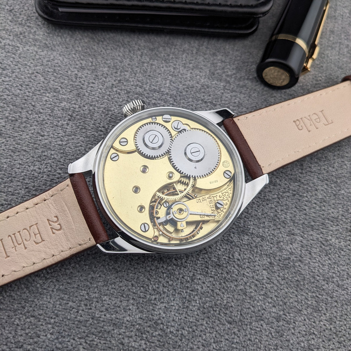 Vista posteriore dell'orologio Omega con retro trasparente, movimento dorato, ingranaggi visibili e cinturino marrone su tessuto grigio.