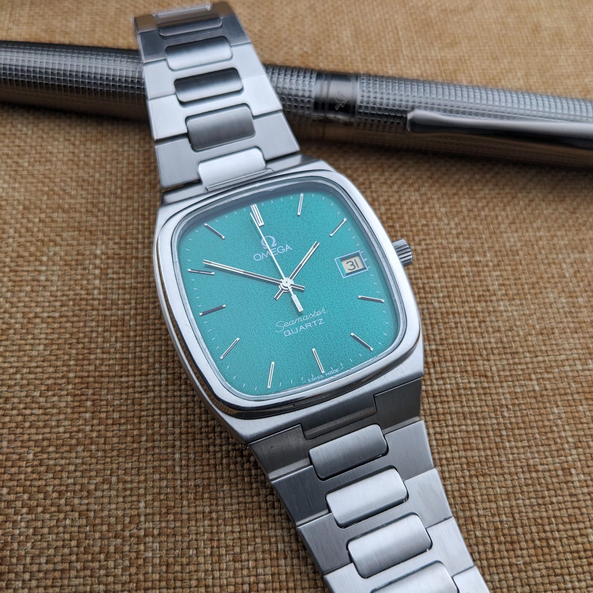 Close angled view of the Omega Seamaster reference 196.0166 teal dial and silver bracelet, positioned beneath a textured black pen.