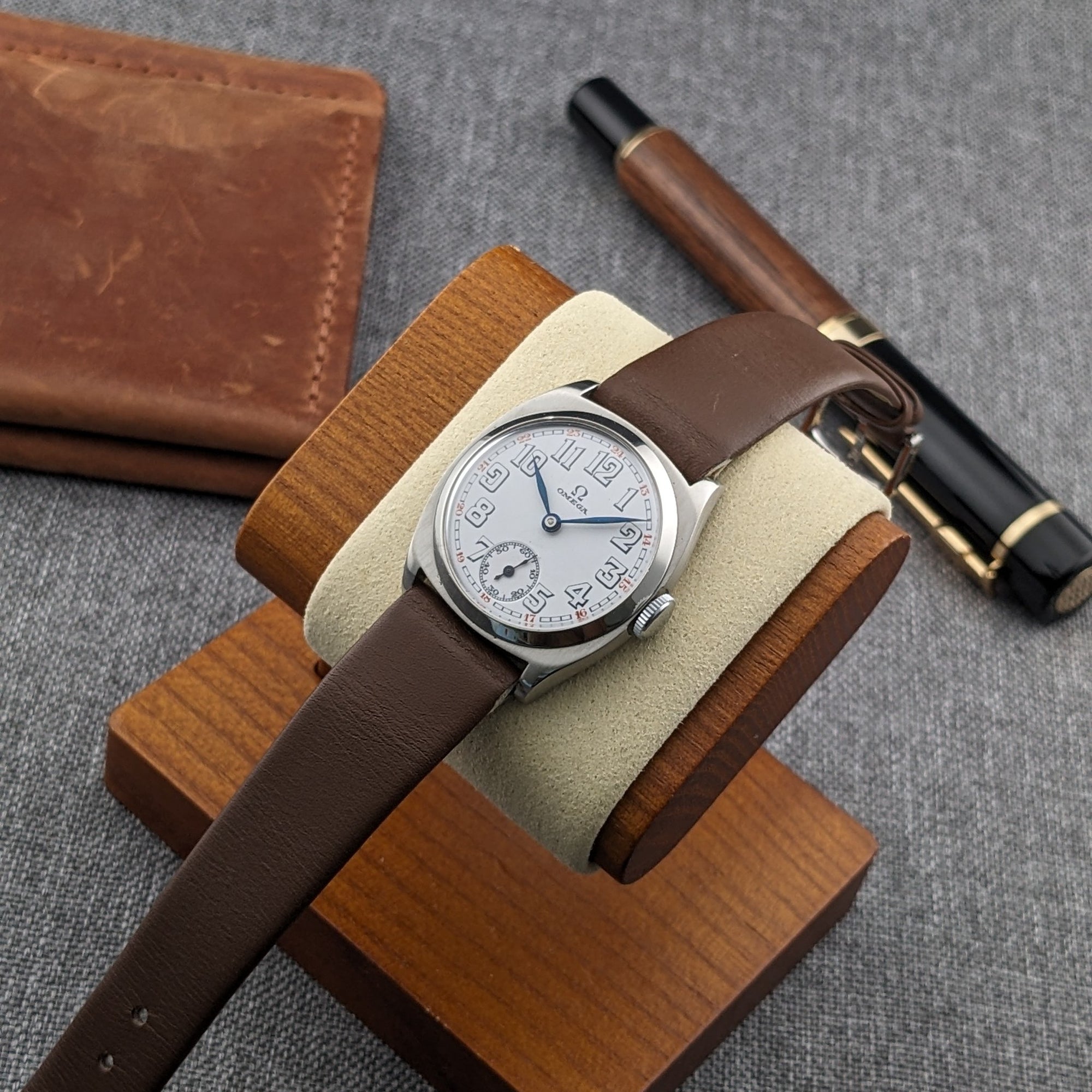 Omega watch resting on a wooden stand with a cream cushion, accompanied by a brown wallet and black-and-wood pen on gray fabric.
