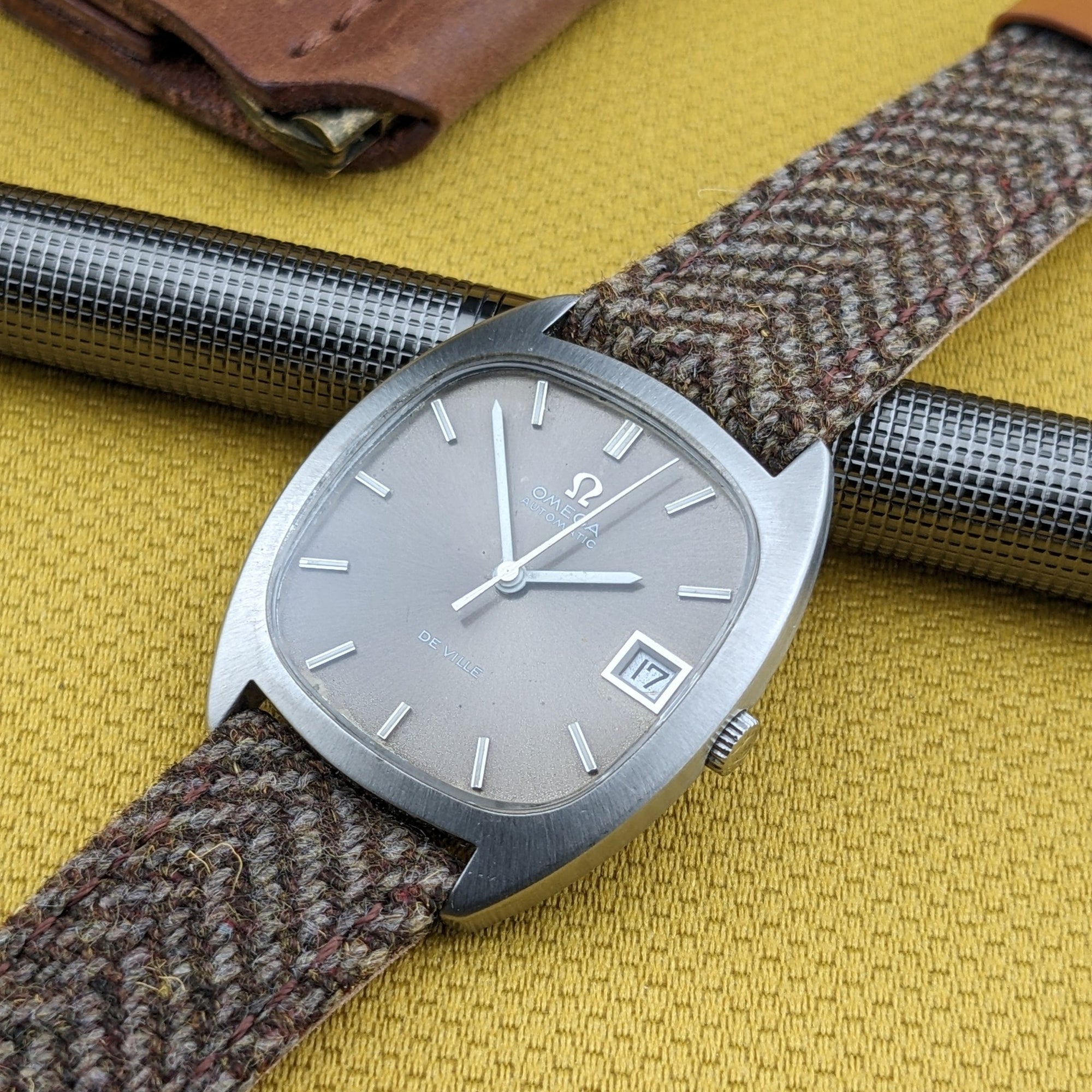 Omega Dr Ville Automatic ref. 162.045 rests diagonally over a metal pen, displaying its silver dial, date window, and textured strap.