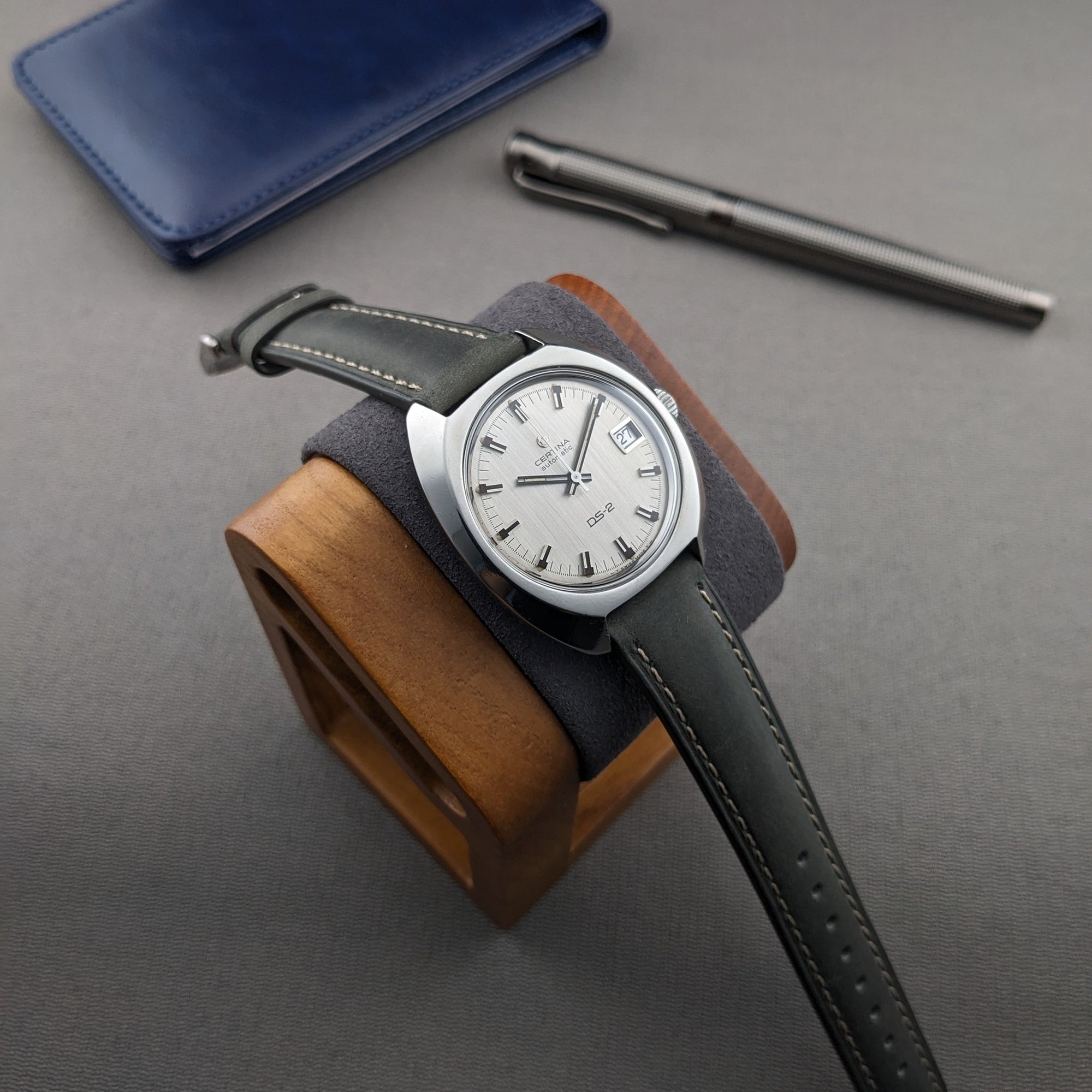 Certina DS-2 Automatic reference 5801 300 displayed on a wooden stand with a blue wallet and metal pen on a gray surface.