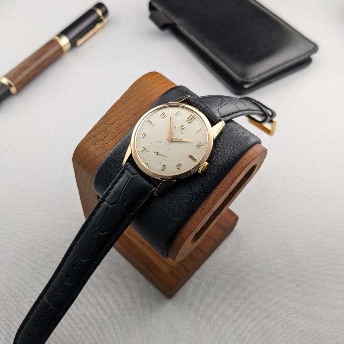 Omega Classic reference 14391-1 resting on a wooden watch stand, with its black strap extending across the foreground beside a pen and case.