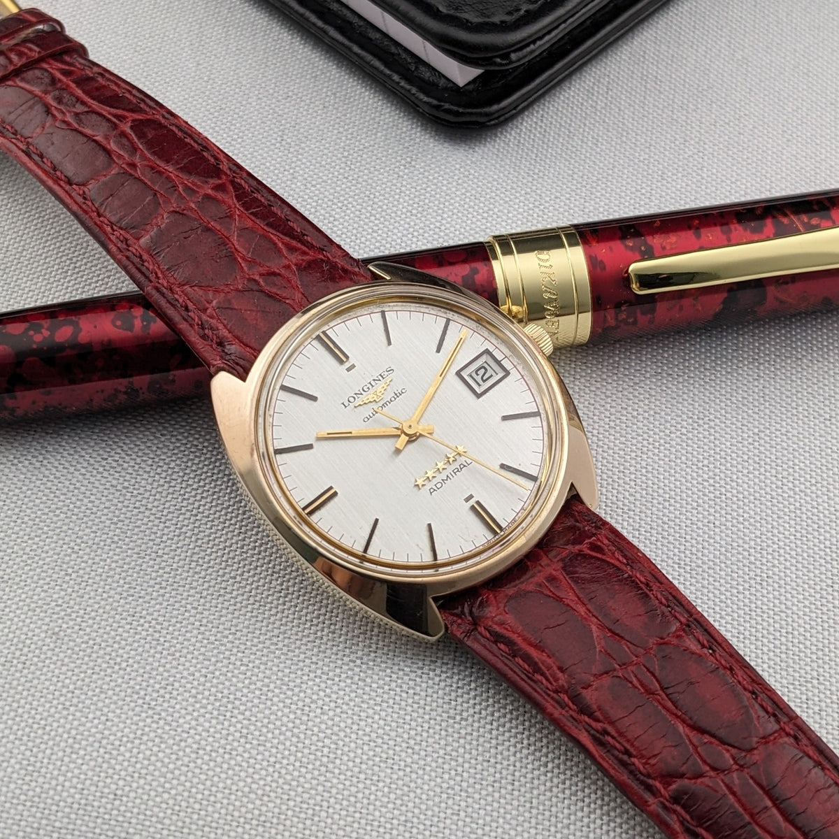 Longines Admiral Referenz 8343-1 diagonal über einem burgunderroten Stift, mit strukturiertem burgunderrotem Armband über den Rahmen.