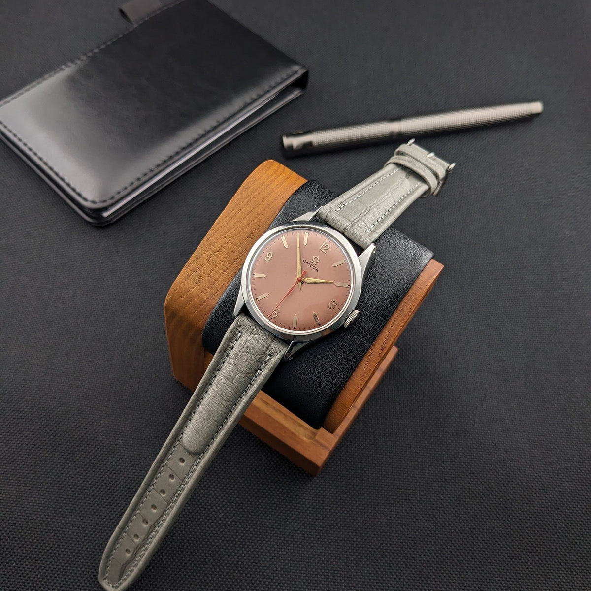 Omega Classic 2760-6 SC auf einem Holzuhrenständer mit ausgezogenem grauem Armband, neben schwarzem Portemonnaie und strukturiertem Metallstift.