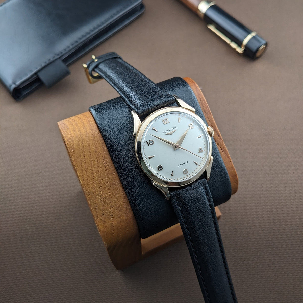Longines Automatik Referenz 6058 auf einem Holz-Uhrenständer, mit schwarzem Portemonnaie und Stift im Hintergrund.
