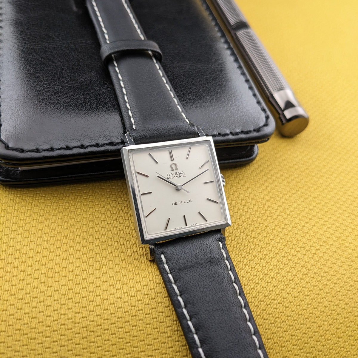 Omega De Ville Automatik 161.014 auf schwarzem Gehäuse, zeigt helles Zifferblatt, quadratisches Gehäuse und schwarzes Armband auf senffarbenem Stoff.