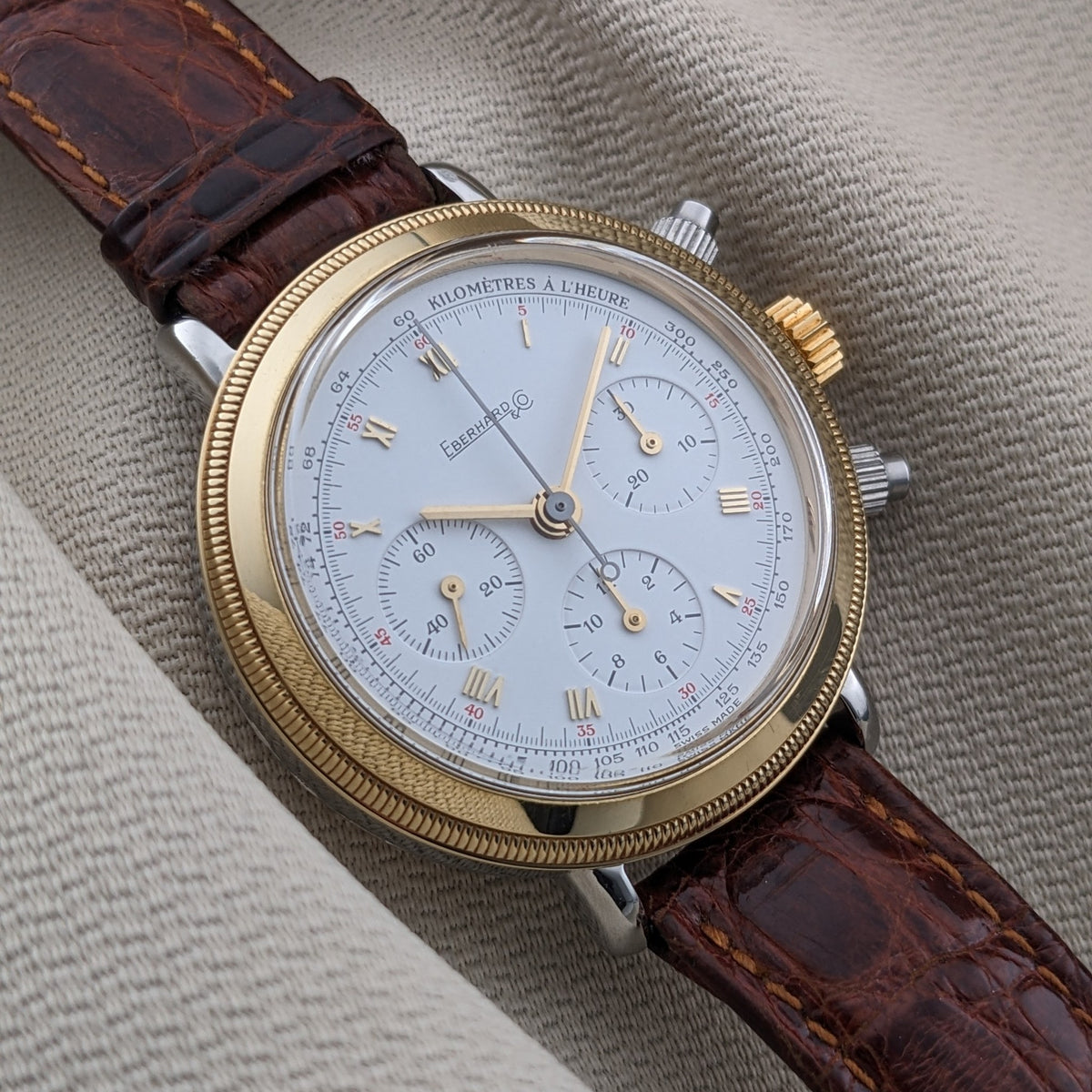 Eberhard & Co.. Godron Chrono reference 32031 shown at an angle on ribbed fabric, highlighting the white dial, gold-tone bezel and brown strap.