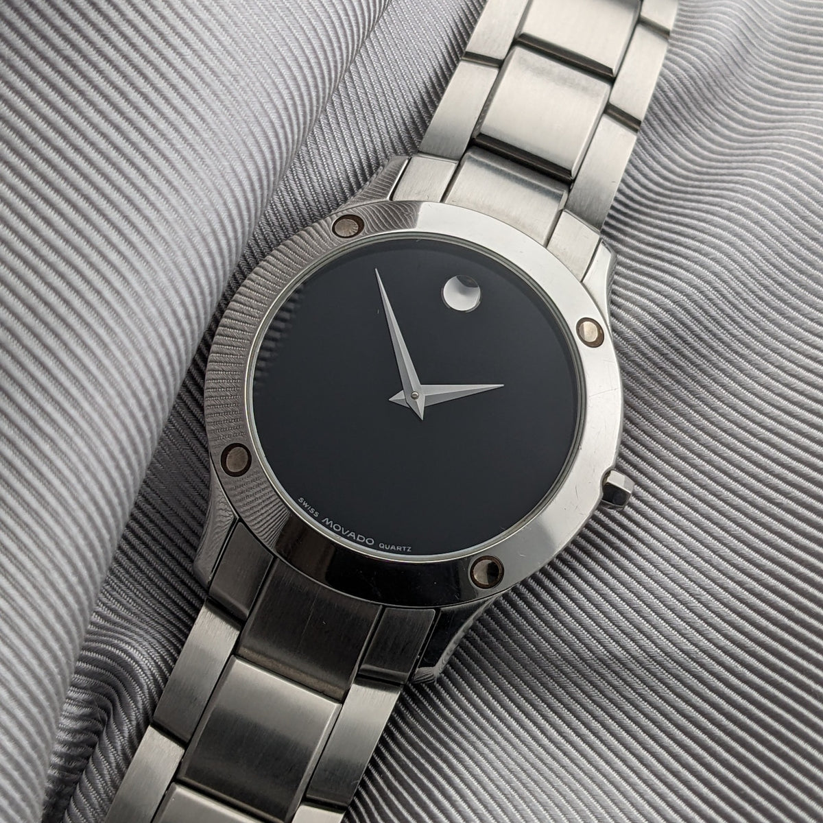 Montre Movado reposant en diagonale sur un tissu côtelé, avec cadran noir, aiguilles argentées, repère circulaire et bracelet en métal.
