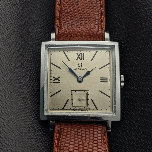 Straight-on Omega Tank dial view shows Roman numerals, blue hands, a rectangular lower aperture, and brown strap against black fabric.