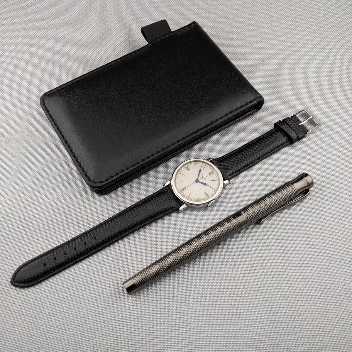 Omega watch laid flat with its long black strap, black wallet and silver textured pen on a gray surface.