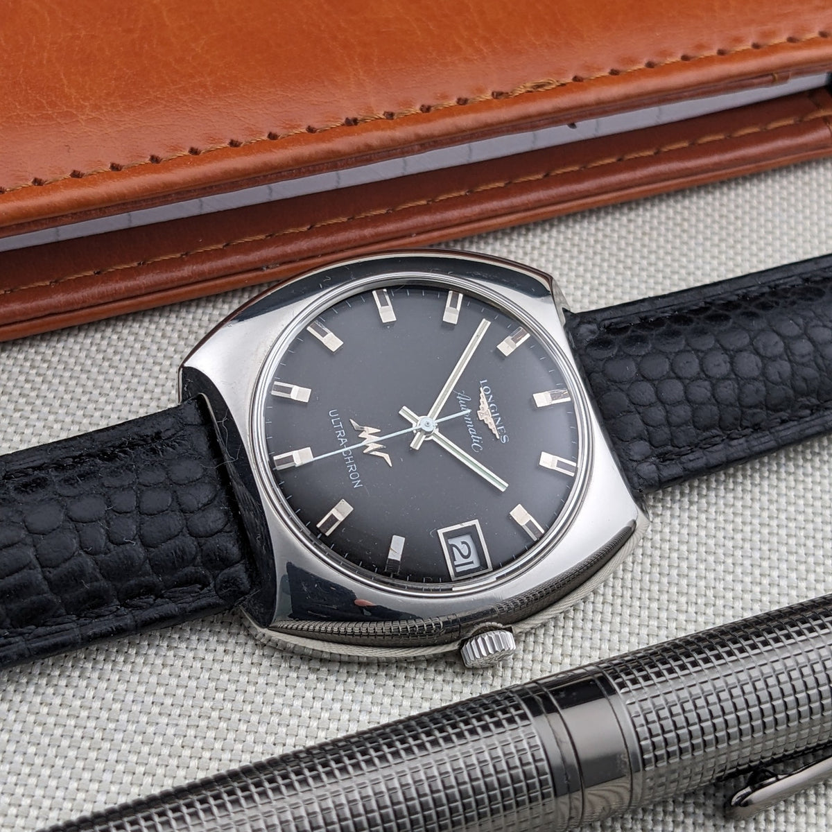 Longines Ultrachron reference 8429 lying diagonally with dark dial, date window, black strap, brown wallet and textured metal pen.