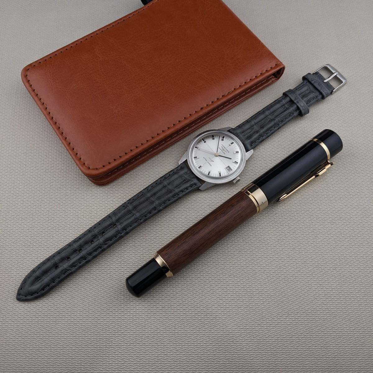 Longines Ultra-Chron 7827 laid flat with its black strap, alongside a wooden-and-black pen and a brown leather case.