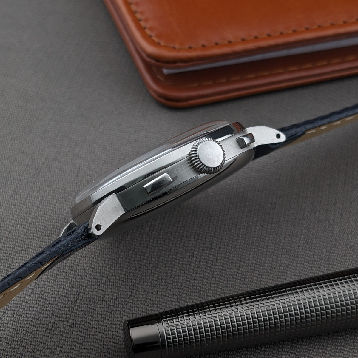 Side profile of the Eberhard & Co. Pre-Extra Fort shows its round case, crown, pushers and blue strap beside a pen.