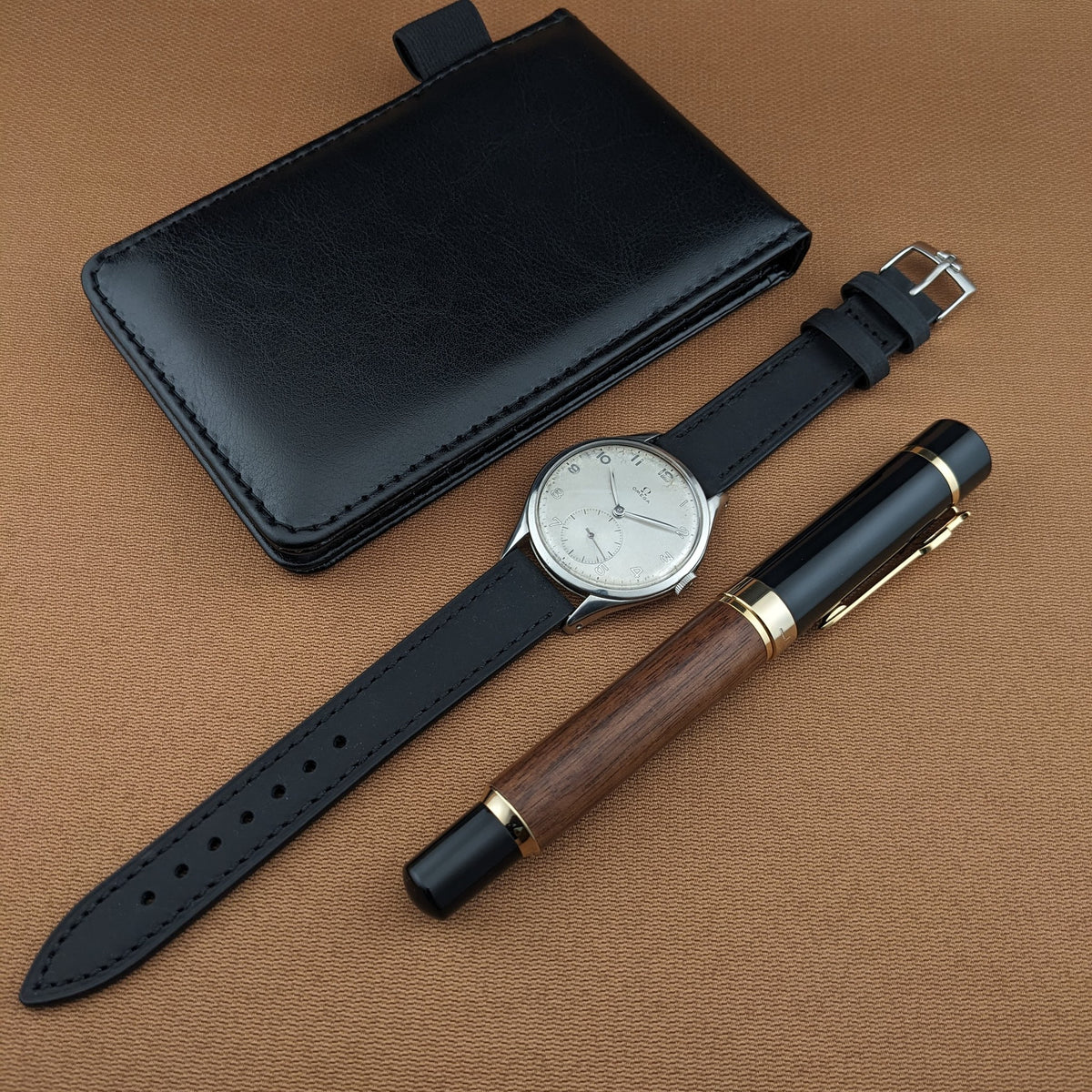 Omega Jumbo reference 2505-5 shown with its black leather strap, black wallet, and wood-and-black pen on brown fabric.