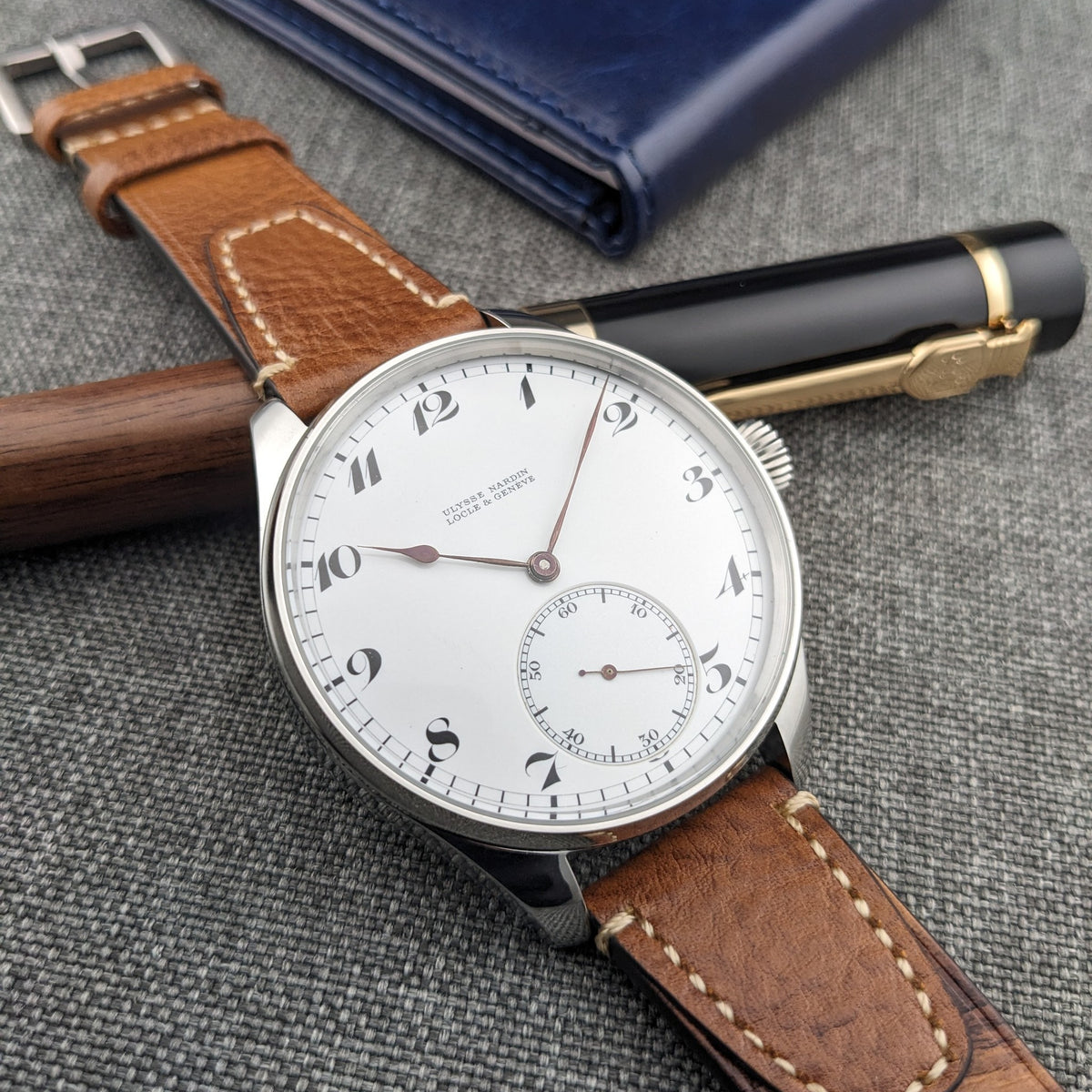 Ulysse Nardin कलाई घड़ी लेखन उपकरणों के पार रखी हुई, जिसमें सफेद डायल, काले अंक, छोटे सेकंड सबडायल और चमड़े का पट्टा दिखता है।