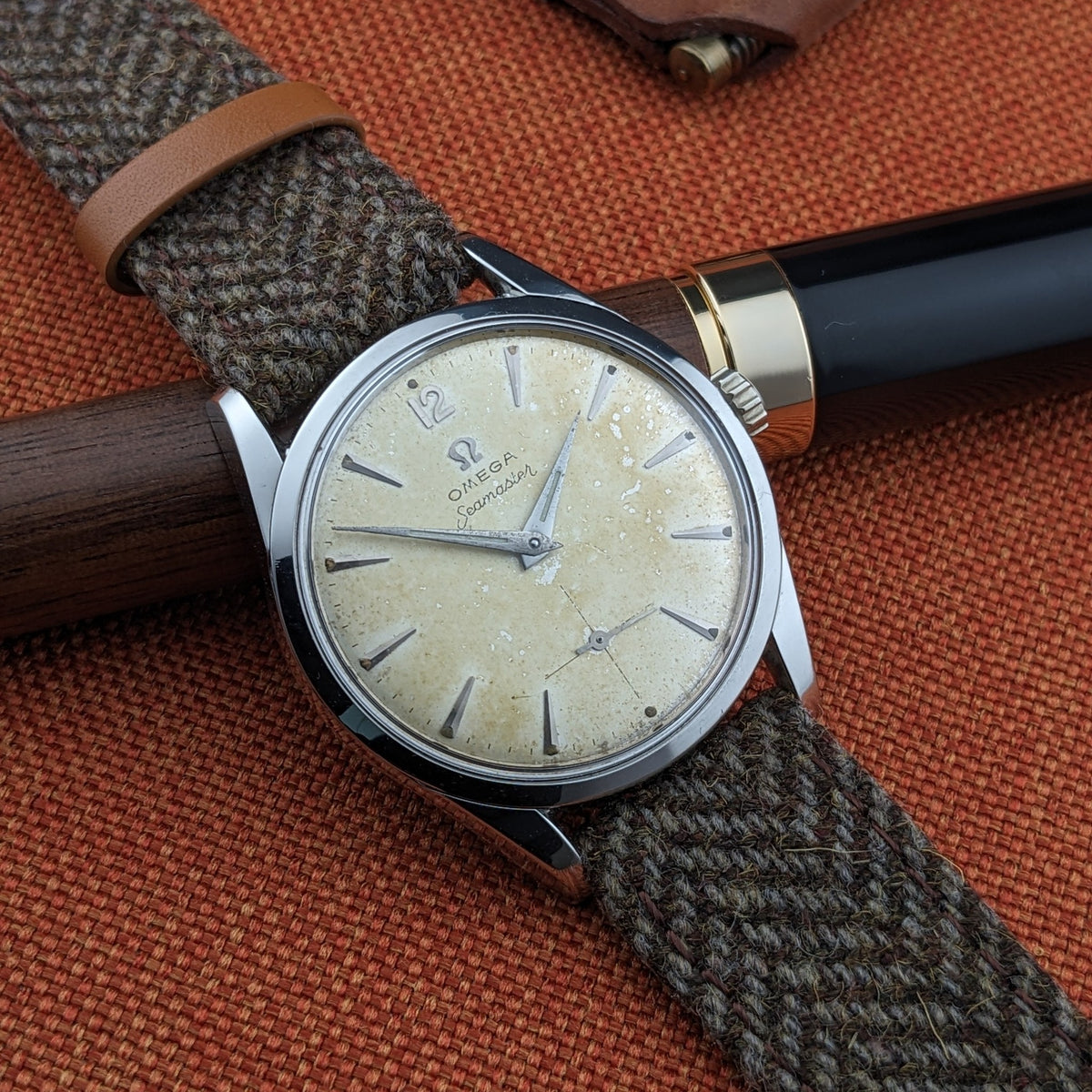 Angled Omega Seamaster reference 29374 beside a black-and-gold pen, showing its cream dial, applied markers and herringbone-style strap.